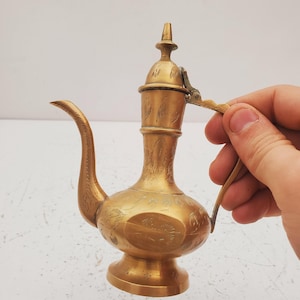 Puede incluir: Una jarra de latón vintage con un diseño ornamentado. La jarra tiene un pico largo y curvo, una tapa con bisagras y un asa. El cuerpo de la jarra está decorado con patrones florales grabados. La jarra es de color dorado cálido.