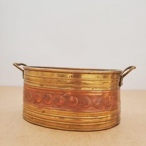 Peut inclure: Jardinière ovale en laiton avec une bande de cuivre. La jardinière est dotée de deux petites poignées et d'une bande décorative de cercles en relief. Le laiton a une teinte dorée chaude, tandis que la bande de cuivre ajoute une couleur brun-rougeâtre contrastante.