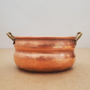 Puede incluir: Una olla redonda de cobre con un acabado metálico pulido y una textura ligeramente martillada. El recipiente cuenta con dos pequeñas asas de latón curvadas unidas a los lados opuestos del borde, creando un contraste de color y material.