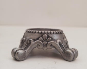May include: A silver-colored, decorative pedestal base with ornate, curved legs. The base has a detailed, textured rim and a floral design. The overall style is reminiscent of antique furniture.