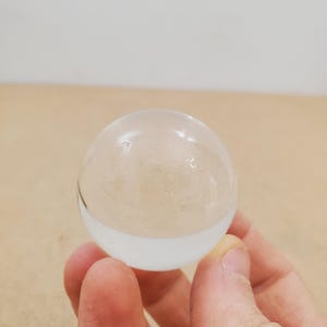 Puede incluir: Una esfera de vidrio transparente con una base blanca, sostenida en una mano. La esfera es clara, permitiendo el paso de la luz. El fondo es una superficie marrón claro y una pared blanca.