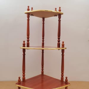 May include: A three-tiered corner shelf with a rich, reddish-brown finish. Each triangular shelf is trimmed with gold-colored accents. The shelf has a classic design, with turned wooden supports and decorative finials.