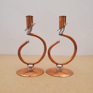 Peut inclure: Une paire de bougeoirs assortis avec une structure circulaire en cuivre, une base plate circulaire en cuivre et des accents argentés. Chaque bougeoir possède une coupelle cylindrique verticale en cuivre soutenue par un bras incurvé.