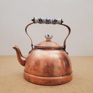 Puede incluir: Una tetera de cobre con cuerpo redondeado y pico curvo. La tetera cuenta con un asa de cerámica blanca y un pomo de tapa a juego, ambos decorados con un patrón de hojas azules. La superficie de metal tiene un acabado pulido y brillante.