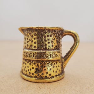 May include: A small, gold-colored miniature pitcher with a handle and spout. The pitcher has a textured surface with small, evenly spaced holes. The word "COCKINGTON" is inscribed on a band around the middle of the pitcher.