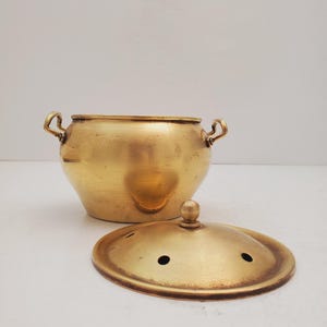 May include: A brass pot with two handles and a lid. The pot has a rounded shape and the lid has a knob on top. The pot and lid have a shiny finish.