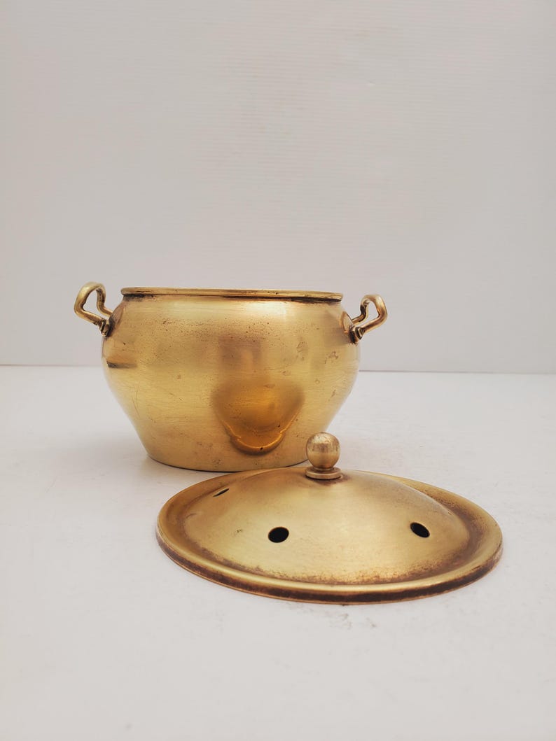 May include: A brass pot with two handles and a lid. The pot has a rounded shape and the lid has a knob on top. The pot and lid have a shiny finish.