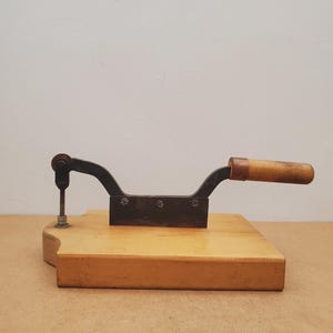 Puede incluir: Una herramienta antigua de madera y metal, probablemente para cortar o dar forma. Cuenta con una base de madera, una cuchilla de metal oscuro y un mango curvo con agarre de madera. La herramienta está diseñada para uso manual.