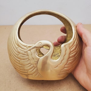 May include: A polished brass bowl shaped like a swan with wings extending upwards to form a circular rim. The metal surface features engraved feather details on the body and wings, with a smooth, curved neck and head facing to the left.