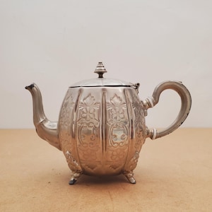 以下が含まれることがあります： 丸みを帯びたボディ、装飾的な花の模様、湾曲したハンドルを備えた銀色のティーポット。 ティーポットには注ぎ口、装飾的なつまみ付きの蓋があり、3つの小さな足の上に置かれています。 ティーポットは金属製です。