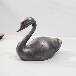 Puede incluir: Figura de cisne plateado con cuello curvo y pico detallado. El cisne tiene un acabado metálico liso y varios agujeros pequeños en la espalda. La figura descansa sobre una superficie blanca.