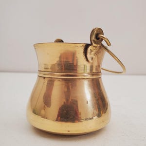 May include: A small, antique brass bucket with a rounded base and a flared rim. It features a curved handle attached to the side. The bucket has a polished, reflective surface, and is likely used for decorative purposes.