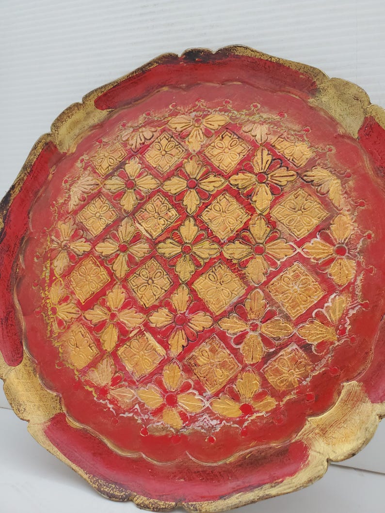 K&ouml;nnte beinhalten: Ein rundes, rotes Holztablett mit einem goldfarbenen Design aus Blumen und geometrischen Formen. Das Tablett hat einen gewellten Rand.