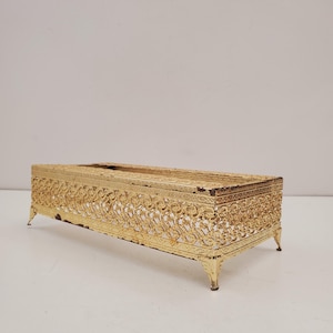 Puede incluir: Una funda rectangular de metal dorado para caja de pañuelos con detalles calados y filigranas ornamentadas. La caja tiene un acabado ligeramente desgastado y descansa sobre cuatro pequeños pies curvos. Diseño vintage y decorativo.