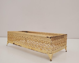 May include: A rectangular, gold-colored metal tissue box cover with ornate, openwork filigree detailing. The box has a slightly distressed finish and rests on four small, curved feet. The design is vintage and decorative.