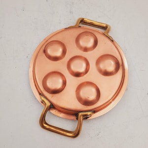 Puede incluir: Una fuente de cobre para servir caracoles con seis huecos. La fuente redonda tiene dos asas de latón y un acabado pulido. El cobre tiene un tono marrón rojizo cálido.