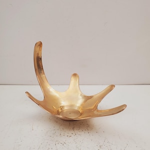 Op de afbeelding: Een goudkleurige decoratieve kom met een geweivormig ontwerp. De kom heeft een centrale cirkelvormige basis en verschillende gebogen, puntige verlengingen. De kom is gemaakt van metaal en heeft een glanzende afwerking.