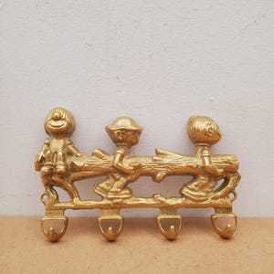 May include: A vintage brass wall hook featuring three cartoon figures holding onto a log. The hook has four curved hooks below the figures. The figures and log are a warm gold color, creating a whimsical and decorative piece.