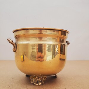 May include: A polished brass bowl with decorative handles and feet. The bowl has a rounded shape and a wide opening. The brass reflects light, creating a warm appearance. This vintage piece is suitable for decorative purposes.