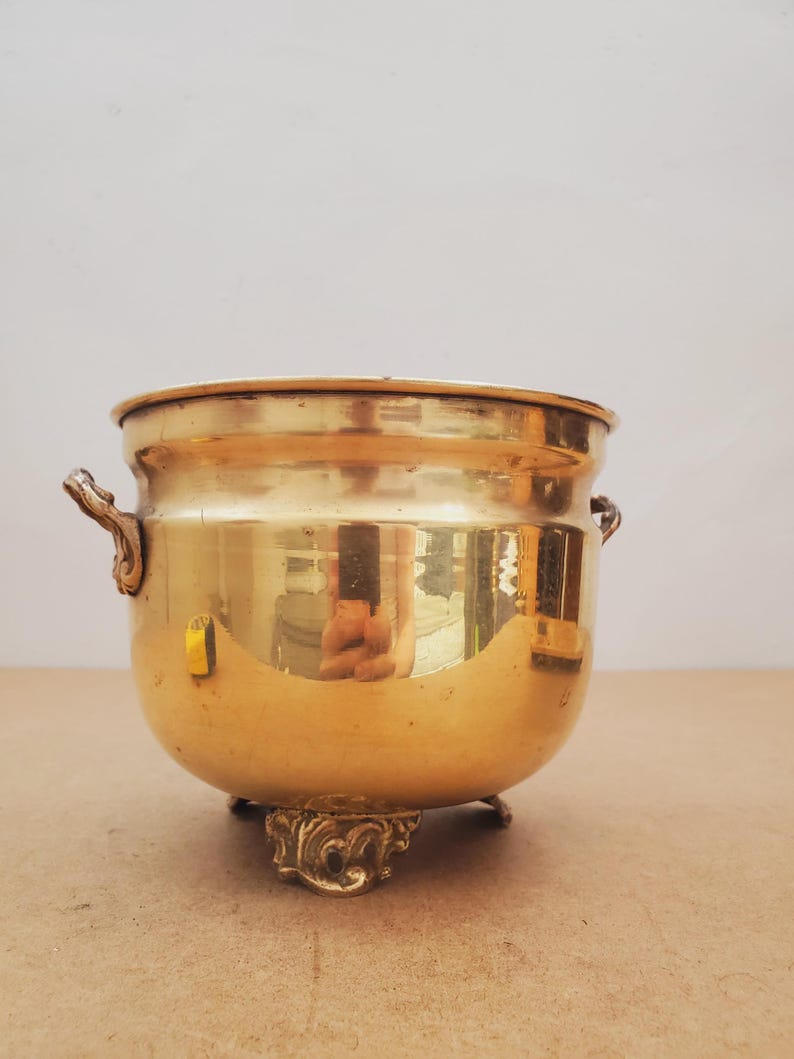 May include: A polished brass bowl with decorative handles and feet. The bowl has a rounded shape and a wide opening. The brass reflects light, creating a warm appearance. This vintage piece is suitable for decorative purposes.
