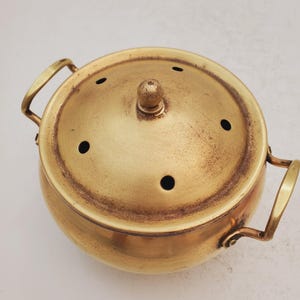May include: A gold metal incense burner with a round body and a lid with small holes. The burner has two handles on the sides.
