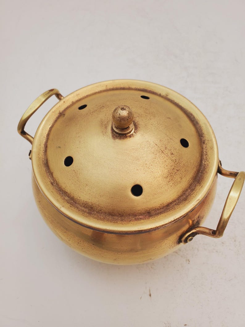 May include: A gold metal incense burner with a round body and a lid with small holes. The burner has two handles on the sides.