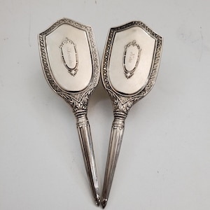 May include: A pair of antique silver-coloured hand mirrors. Each mirror features a shield-shaped head with intricate detailing and a slender, ridged handle. The heads have an oval inscription. The mirrors are set against a light background.