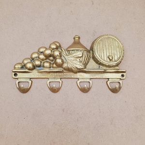 May include: A polished brass wall-mounted hook rack featuring a decorative relief design of a cluster of grapes, a wine bottle, and a wooden barrel. The horizontal bar includes four integrated hooks and two screw holes for mounting on a wall.