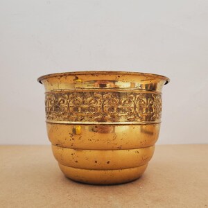 Peut inclure: Un pot en métal doré avec une bande décorative à motifs floraux. Le pot a une forme arrondie avec un bord légèrement évasé et une série de bandes horizontales. La surface montre des signes d'usure, ajoutant à son attrait vintage.