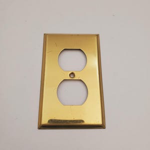 May include: A rectangular, gold-coloured electrical outlet cover with two circular openings and a small hole centred between them. The cover has a slightly bevelled edge and a smooth, reflective surface.