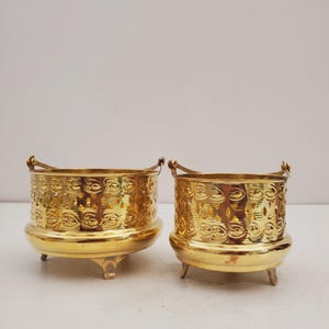 May include: Two gold-colored metal planters with intricate cut-out designs. Each planter has a rounded base, three small feet, and a handle. The planters are likely for decorative purposes, adding a touch of elegance to any space.
