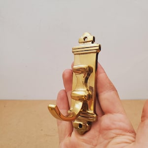 May include: A polished brass wall hook featuring a rectangular backplate with a decorative top and bottom finial, each containing a single screw hole. The hook has a curved, protruding arm extending from the centre of the vertical plate.