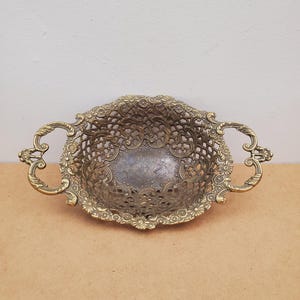Op de afbeelding: Een antieke ovale messing mand met ingewikkelde bloemen- en krulwerkdetails. Het open ontwerp heeft een delicaat patroon en de mand heeft twee sierlijke handvatten. De mand wordt waarschijnlijk gebruikt voor decoratieve doeleinden.