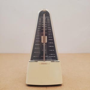 May include: A cream-coloured Seiko metronome with a clear plastic cover. The metronome features a black and white tempo scale and a central pendulum. The tempo markings are visible. The base of the metronome has the Seiko logo. The metronome measures approximately 21 cm in height.
