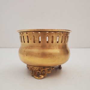 May include: A vintage brass bowl with a decorative base. The bowl has a rounded body and a perforated rim. The base features ornate, scrolled detailing. The brass has a warm, golden hue, suggesting age and use. The bowl is likely a decorative or functional item.