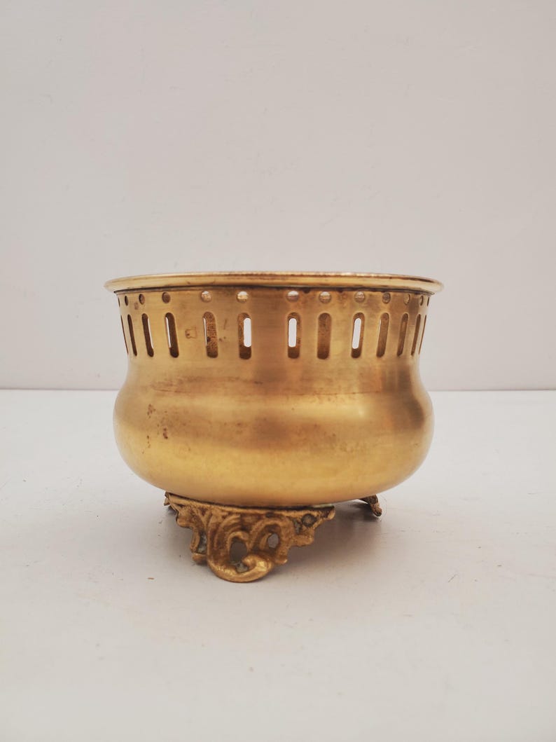 May include: A vintage brass bowl with a decorative base. The bowl has a rounded body and a perforated rim. The base features ornate, scrolled detailing. The brass has a warm, golden hue, suggesting age and use. The bowl is likely a decorative or functional item.