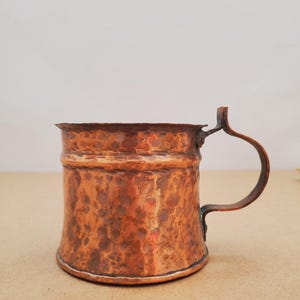 May include: A copper mug with a hammered finish and a curved handle. The mug is cylindrical with a flared rim. The copper has a reddish-brown hue with darker patches. The handle is attached to the side. The mug is likely for drinking.