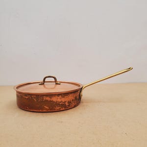 May include: A round copper sauté pan with a matching lid and a long brass handle. The exterior of the copper pan shows signs of wear and patina, while the lid features a small, arched brass handle attached to the centre of the flat surface.