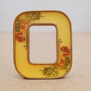 May include: A rectangular picture frame with rounded corners, featuring a yellow background and a floral pattern of orange roses and small white flowers. The frame has a metallic gold-toned border that outlines both the outer and inner edges.