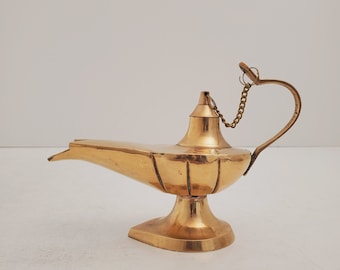 Könnte beinhalten: Eine goldfarbene Dschinnenlampe mit einem dekorativen Griff und einer Kette. Die Lampe hat einen Ausguss und einen breiten Standfuß.