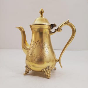Puede incluir: Una tetera de metal de color dorado con un diseño adornado y tres patas. La tetera tiene una tapa con un pomo en la parte superior.