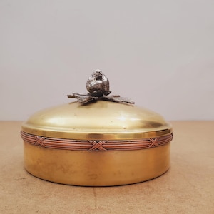 May include: A round, brass-colored container with a lid. The lid is topped with a silver-colored, textured ornament. A copper-colored band with an X-shaped pattern encircles the container. The container sits on a light brown surface.