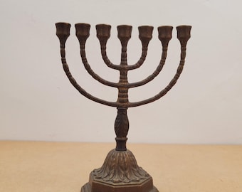 Può includere: Un menorah color bronzo con sette portacandele. La base è di forma ottagonale con dettagli decorativi. Le braccia si incurvano verso l'alto, ciascuna con un portacandela. Il menorah è un simbolo della fede ebraica.
