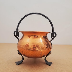 May include: A small, hammered copper cauldron with a twisted black metal handle and three decorative feet. The copper pot has a shiny, reflective surface and is designed with a rounded shape. The handle is attached to the pot with decorative metal brackets.