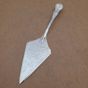 May include: A silver cake server with an ornate floral pattern on the blade and handle. The server has a triangular blade with a serrated edge and a decorative handle. The server is made of a shiny, reflective metal.