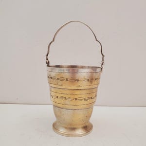 Può includere: Un secchio in metallo argentato con manico curvo. Il secchio ha una fascia color oro al centro con linee orizzontali incise. La parte inferiore è color argento e il manico è attaccato al bordo.