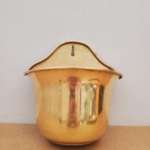 May include: A shiny, gold-colored wall planter with a curved top and a rounded body. The planter has a keyhole for hanging and is made of a reflective material, likely metal. The interior is not visible.