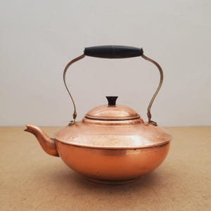 May include: A copper kettle with a black handle and a matching black knob on the lid. The kettle has a rounded body and a curved spout. The handle is attached to the kettle with brass-colored metal supports. The kettle is sitting on a light brown surface.