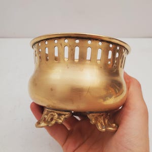 May include: A small, antique brass bowl with a rounded body and three ornate feet. The rim features a series of vertical cutouts. The bowl has a polished, golden finish and is held in a hand.