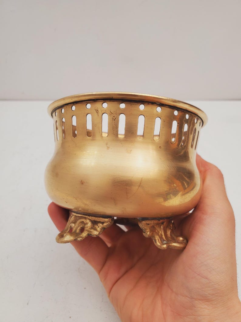 May include: A small, antique brass bowl with a rounded body and three ornate feet. The rim features a series of vertical cutouts. The bowl has a polished, golden finish and is held in a hand.